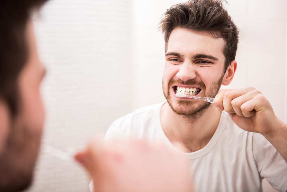 Young man looking in mirror while brushing his teeth 1000x668 - لماذا لا يجب أن تتجاهل الأسنان المكسورة